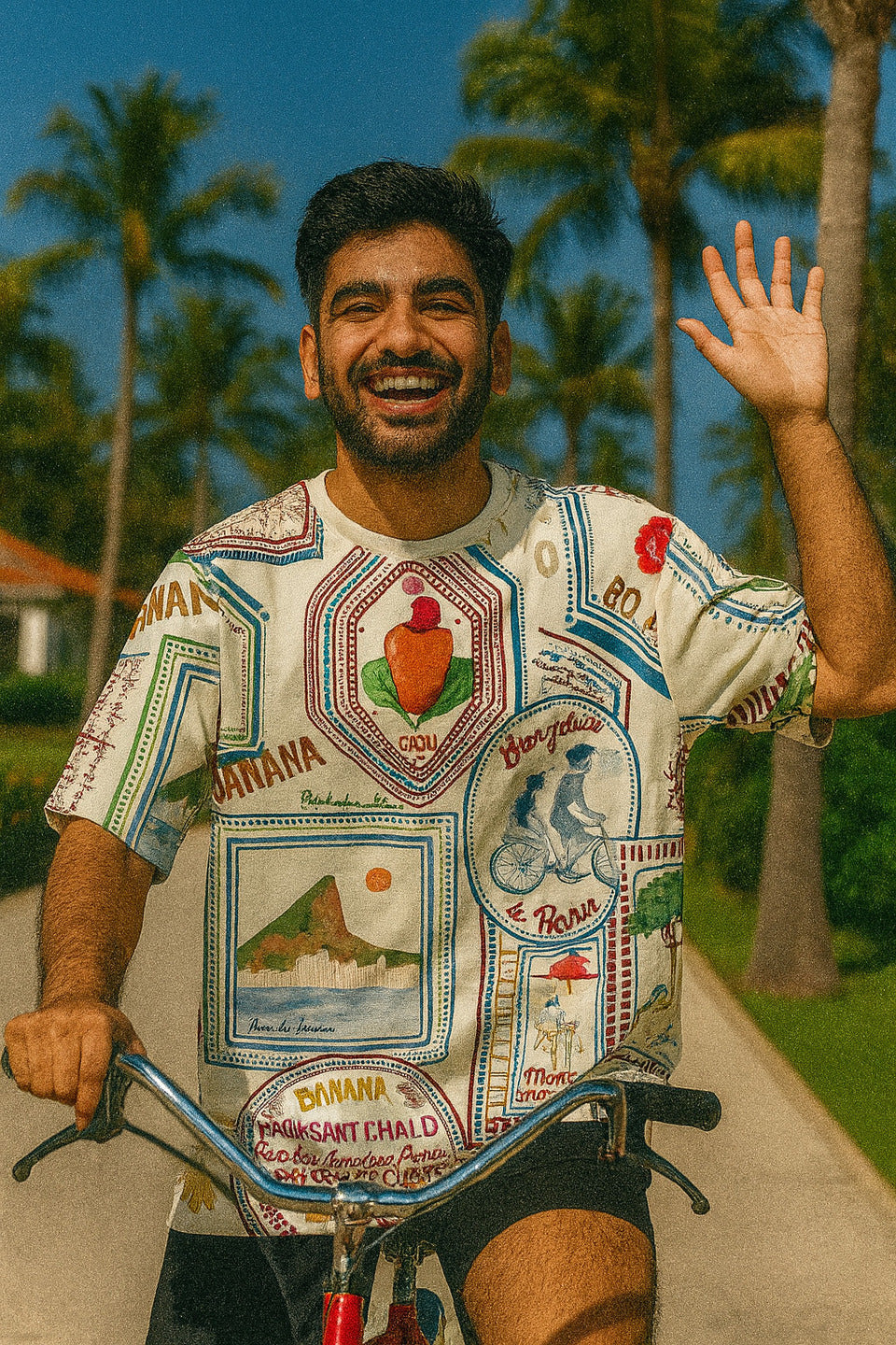 Man with a colorful Bonjour Unisex Oversized T-Shirt From The Edi Archive, and bicycle waving, palm trees in the background