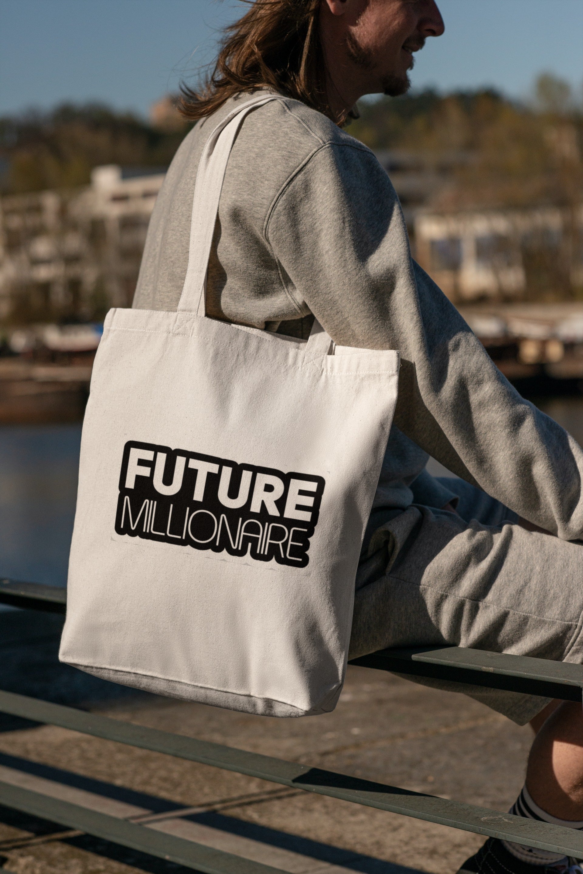 Person holding a tote bag with 'Future Millionaire' text by a waterfront.