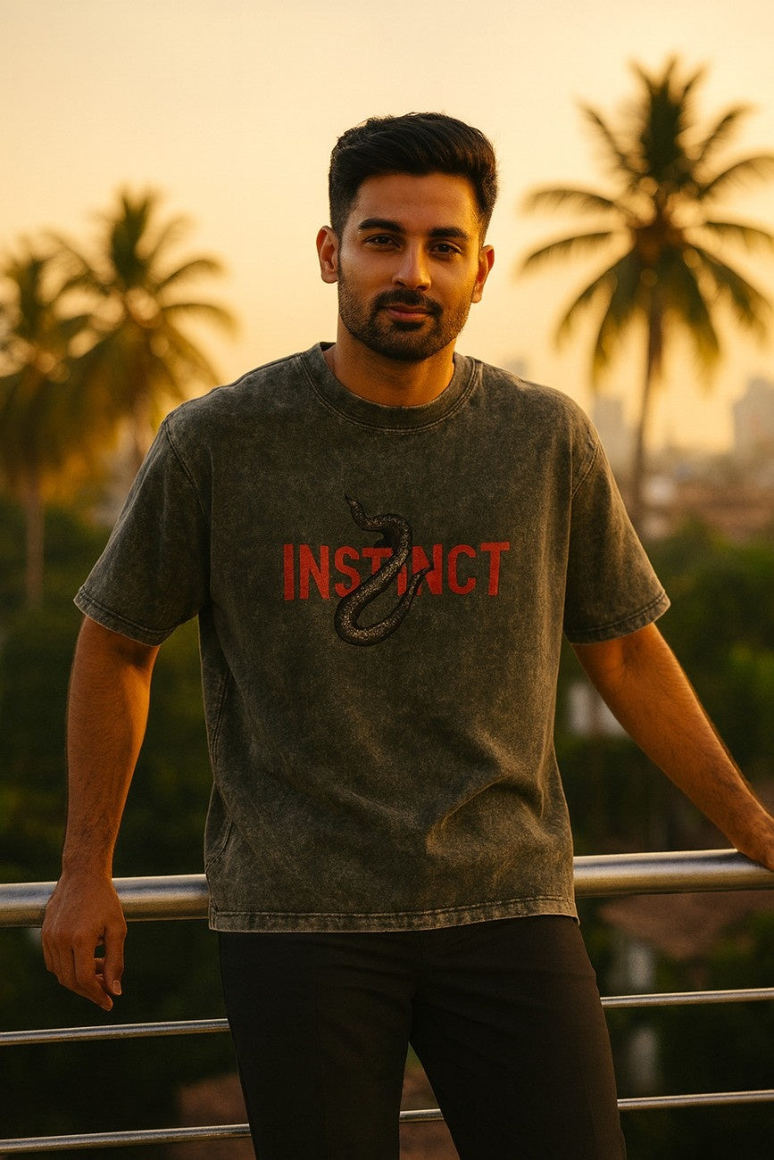 Man wearing an Olive Grey, Instinct Unisex Oversized T-Shirt from The Edi Archive, standing outdoors with palm trees in the background.