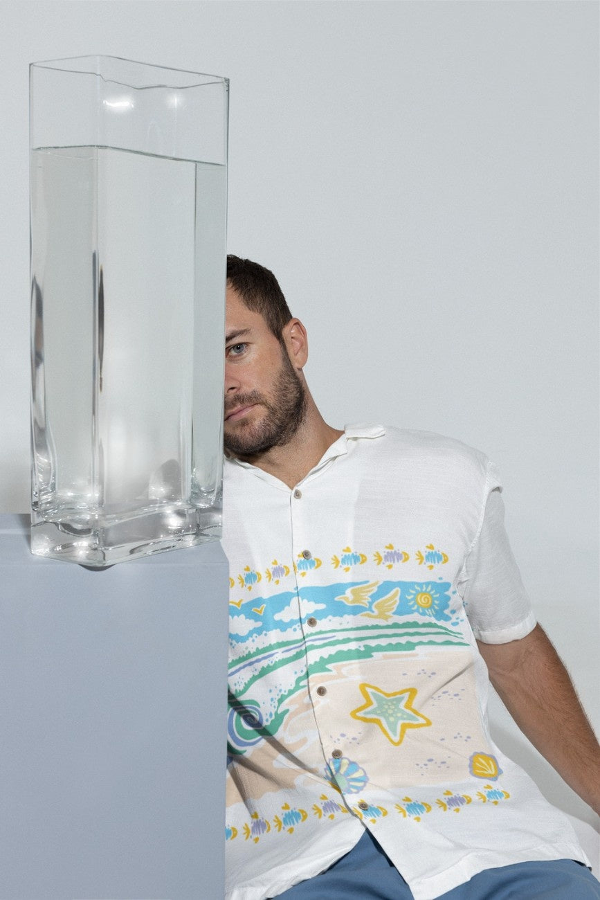 Man wearing a white Beach Bed Men's Hawaiian Shirt from The Tropical Drops Collection from The Edi Archive, peeking from behind a clear vase on a light gray background