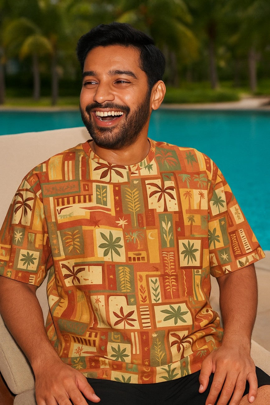 Man wearing a patterned Summer Dreams Unisex Oversized T-Shirt from The Edi Archive, sitting by a pool with palm trees in the background