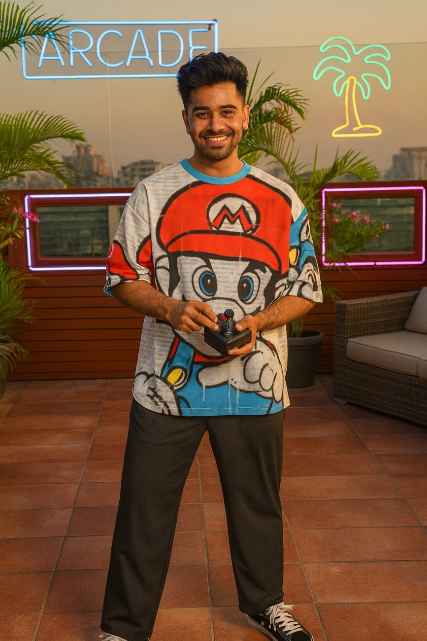 Man wearing a colorful  Super Mario Unisex Oversized T-Shirt From The Edi Archive, standing in an outdoor setting with neon signs and plants.