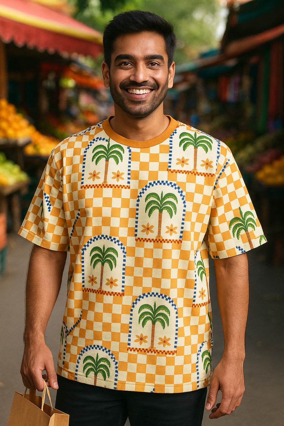 Man wearing a patterned Desert Palm Unisex Oversized T-Shirt from The Edi Archive, with palm trees and checkered design, standing in an outdoor market.