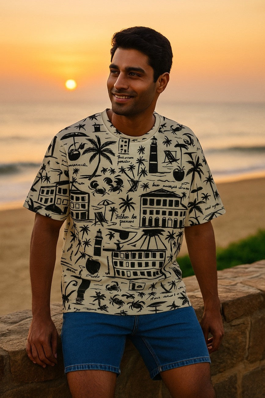 Man wearing a patterned oversized T-Shirt, Live Wild from The Edi Archive and shorts standing on a beach at sunset.