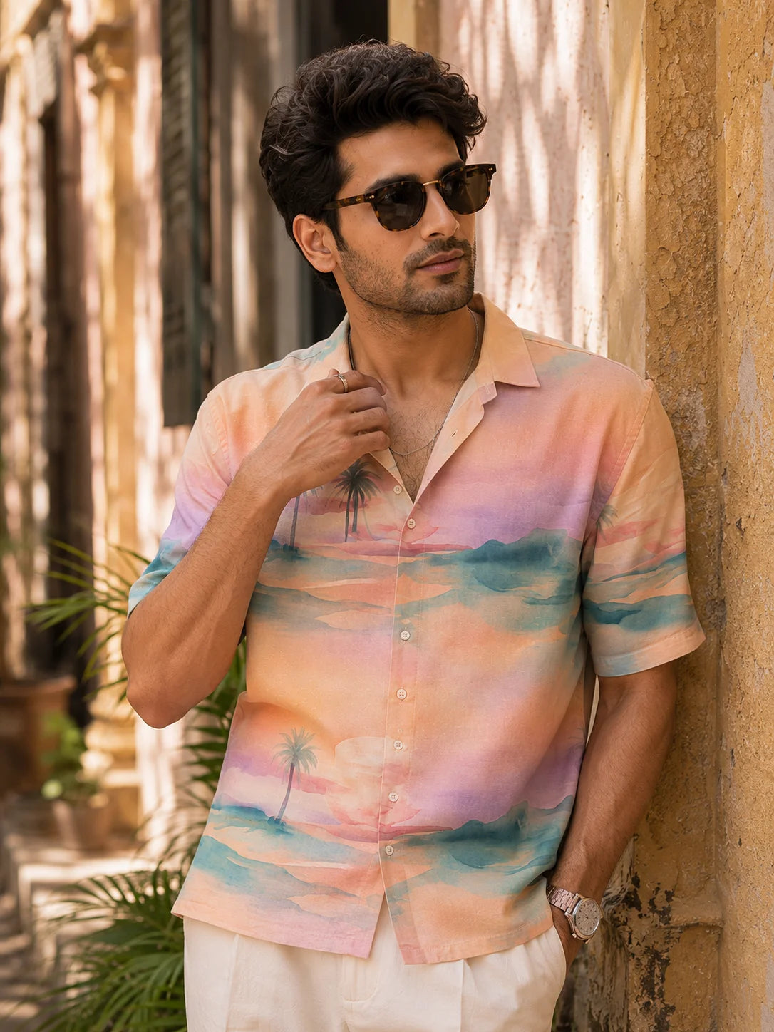 Man in a pastel watercolor sunset print Cuban collar shirt with sunglasses leaning against a sunlit wall, close detail of the cotton linen fabric, Mirage Hawaiian Shirt, Tropical Drop, The Edi Archive